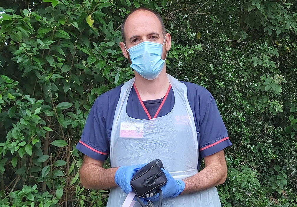 A photograph of Dave on a patient visit. He stands in front of foliage and is wearing PPE. He has a kit bag in his hands.