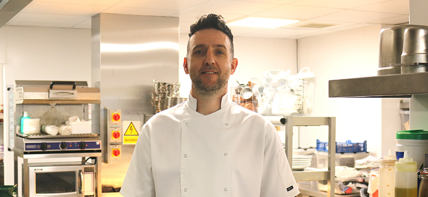 A portrait photo of our chef Gavin Duce. He stands in the kitchens and smiles to the camera.