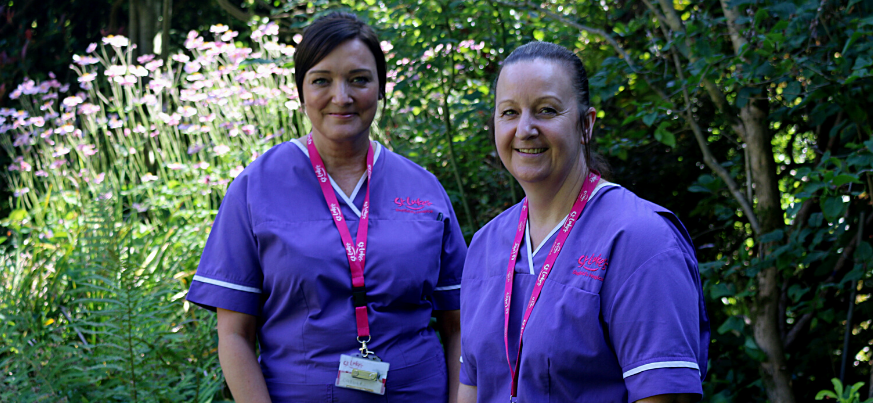 A portrait photograph of Joanne and Catherine. They stand next to each other in the St Luke's gardens and smile to the camera. They are wearing their uniforms.