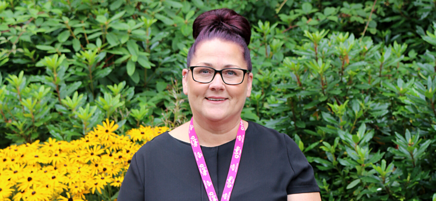 A portrait photograph of Marie Shaw. She is wearing a black top and black-rimmed glasses. She is smiling and stands in front of foliage in the St Luke's gardens.