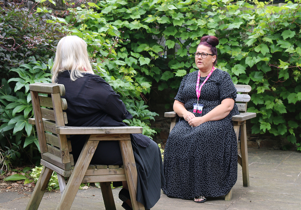 A photograph of Marie Shaw speaking with a client. We can see the client from behind. They sit in a garden. Marie is listening intently.