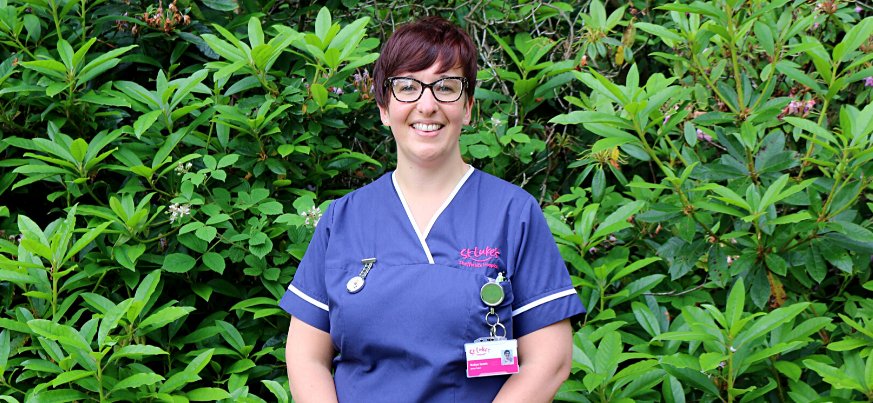 A portrait photo of Robyn Smith, Junior Sister. She is wearing her uniform and smiling to the camera. She stands in front of some greenery in the St Luke's gardens.