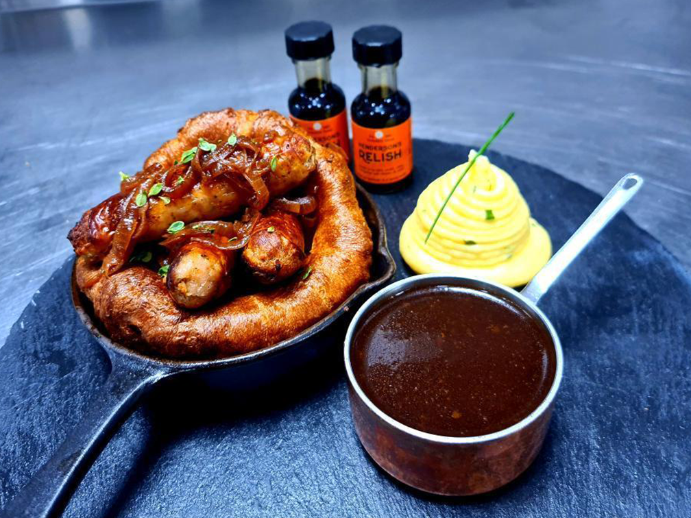 A photograph of Toad in the Hole. There is a pot of gravy next to it, and some small bottles of Henderson&#39;s Relish in the background.